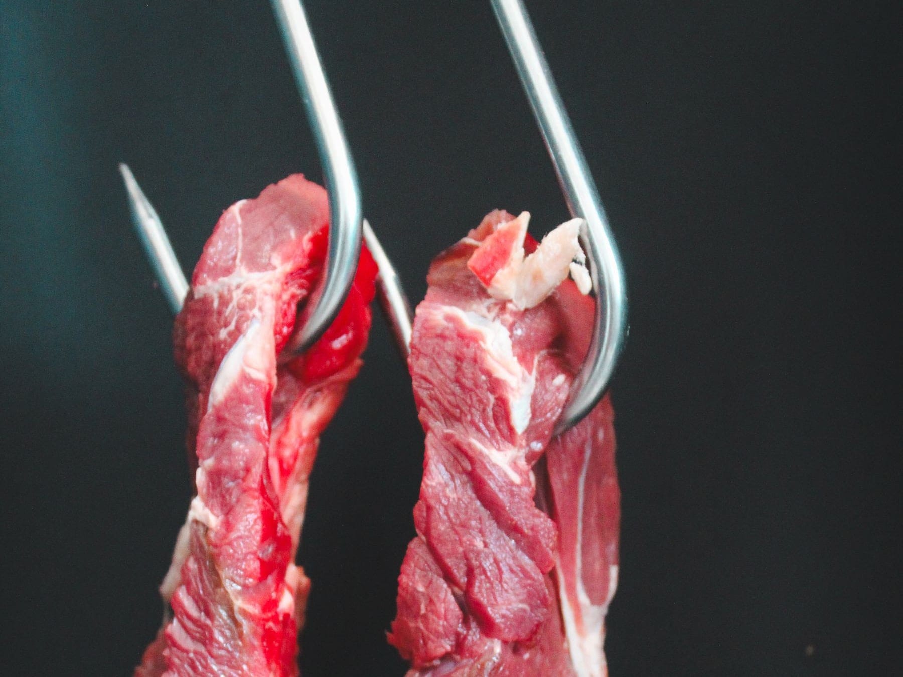 Two cuts of raw beef on steel hooks